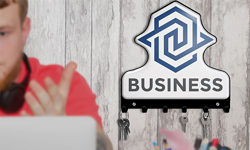 Duine le héide dearg ag ríomhaire glúine, cluasgléas cluaise ar an mhangar; bord eochracha le lógó BUSINESS agus eochracha ar an mballa