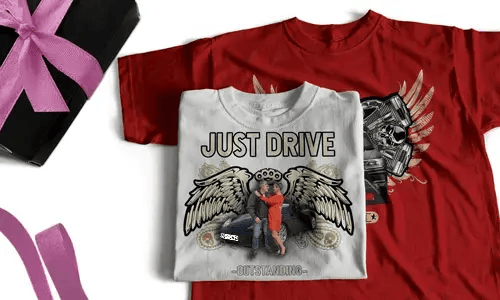 Zwei T-Shirts mit auffälligen Grafiken, eines weiß mit JUST DRIVE und Paar am Auto, eines rot mit geflügeltem Motor und Flammen, neben Geschenkbox mit rosa Schleife